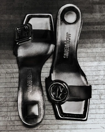 Shiny Jacquemus women's sandals in black and white, presented at an upward angle on a wood-grain surface. Stylish, modern footwear.