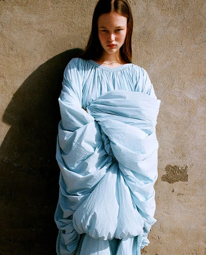 Fashion editorial shot of a young woman wearing a draped light blue dress standing indoors against a tan wall, with natural light.