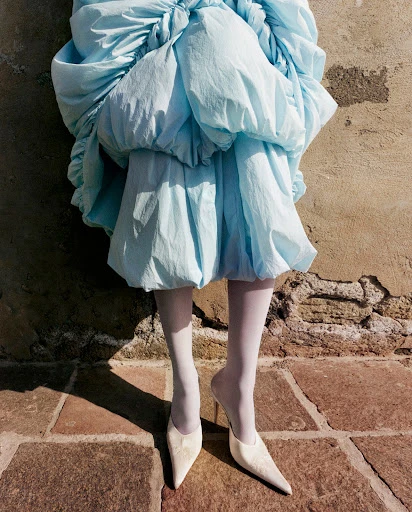 Fashionable woman's legs in white tights and cream-colored heels under a puffy light blue dress. Textured stone wall background and brick paving.