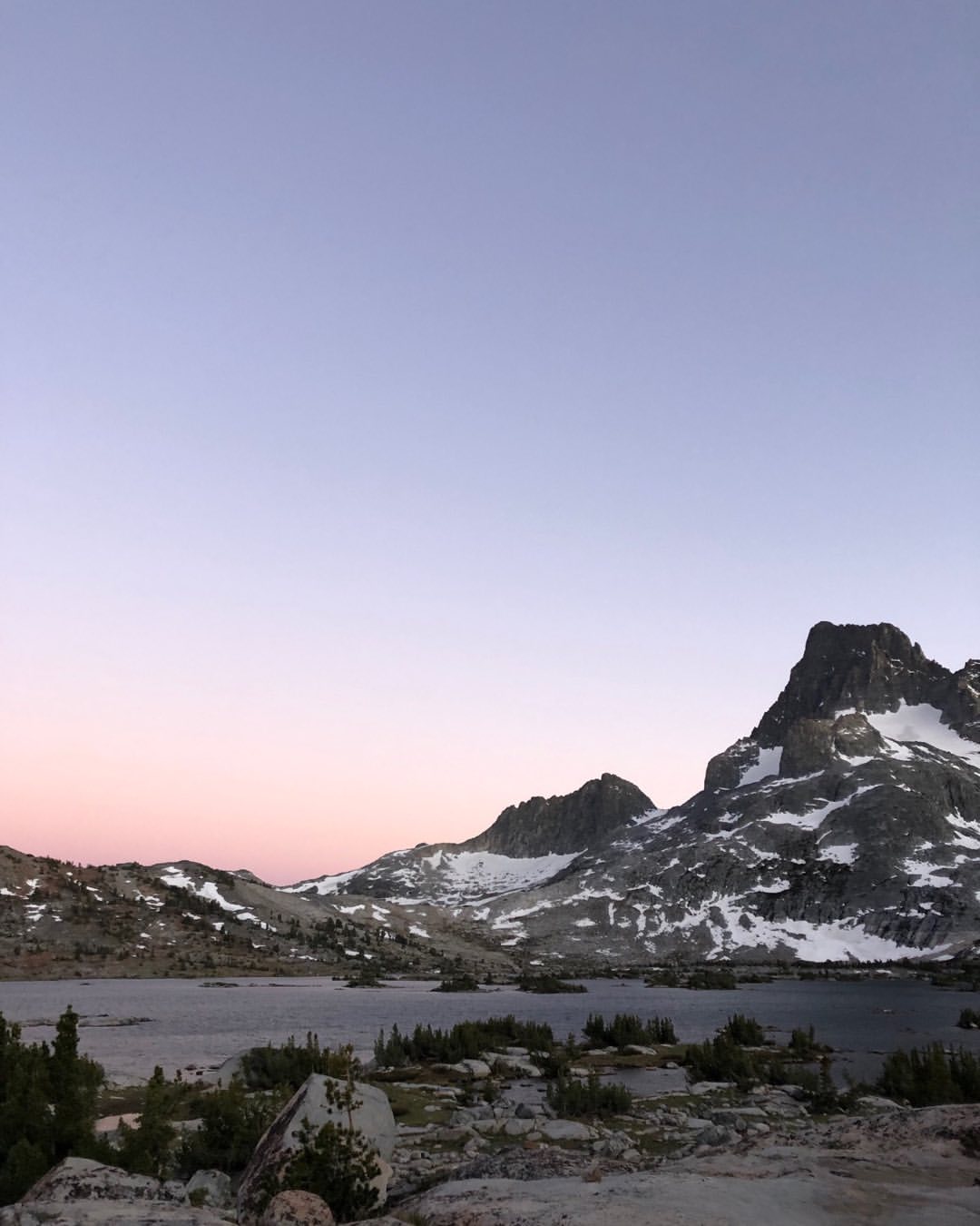 Scenic mountain range landscape view with a lake and snow on the mountain peaks against a blue, pink and purple gradient sky.