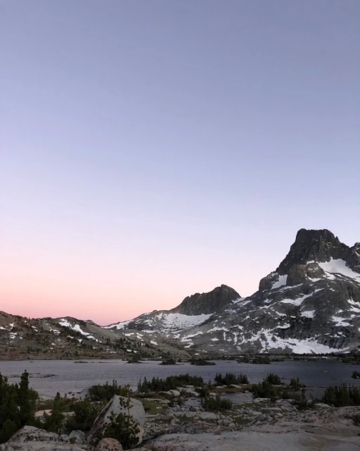 Scenic mountain range landscape view with a lake and snow on the mountain peaks against a blue, pink and purple gradient sky.