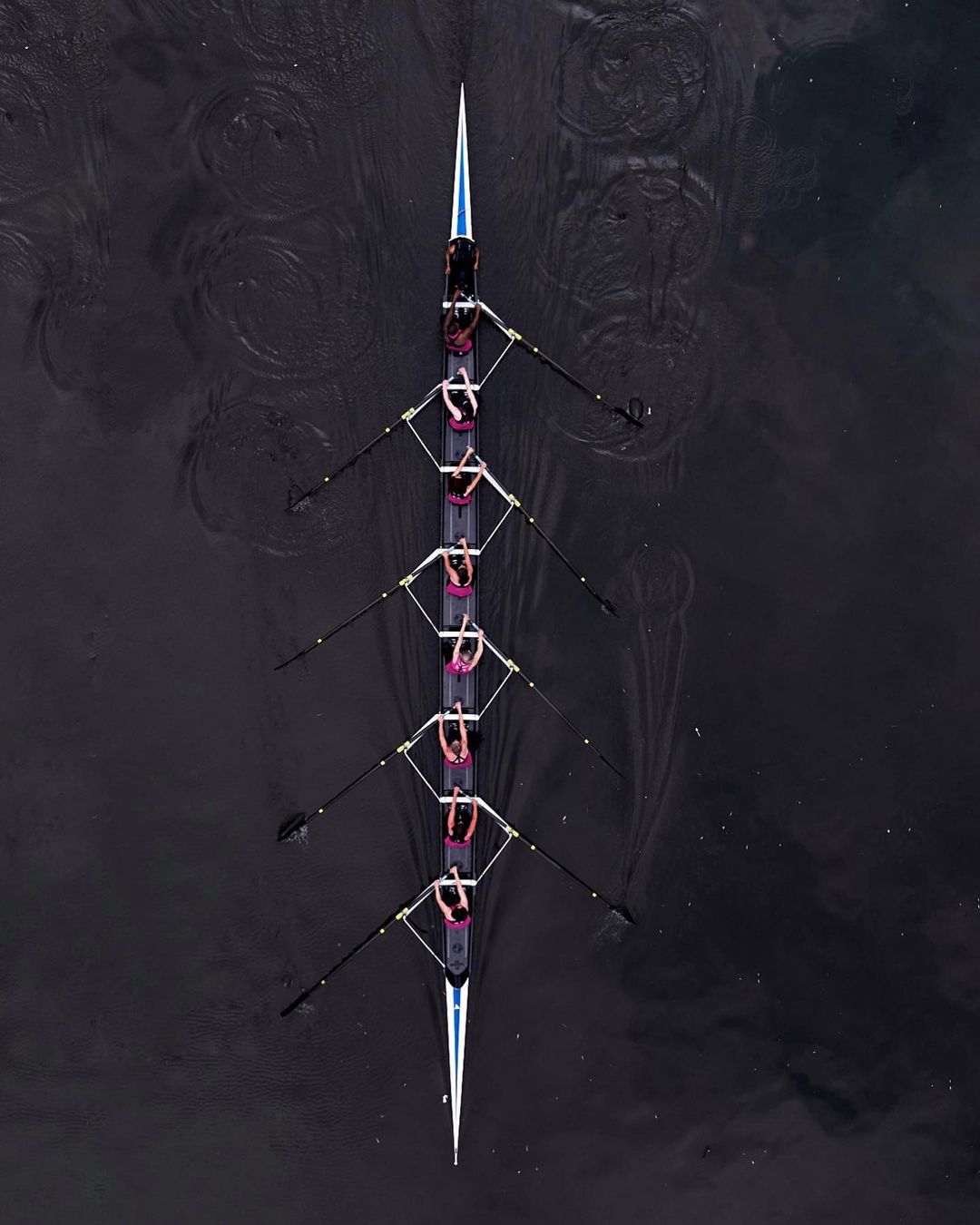 Aerial shot of eight rowers propelling a sleek racing shell across dark water with oars, creating concentric rings on the water's surface.