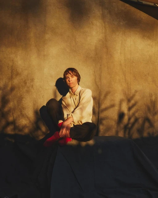 Portrait of a young adult sitting on a draped surface in a photo studio, with warm lighting and shadows of leaves visible on the textured backdrop.