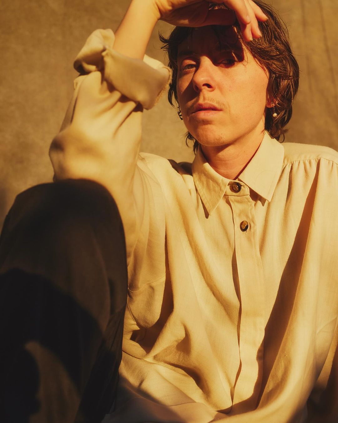 Portrait of a person in a light cream button-down shirt with one hand raised, under warm golden hour lighting