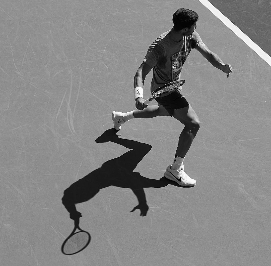 A man is in motion playing tennis on a gray tennis court. He's holding a tennis racket, with his shadow visible on the court.