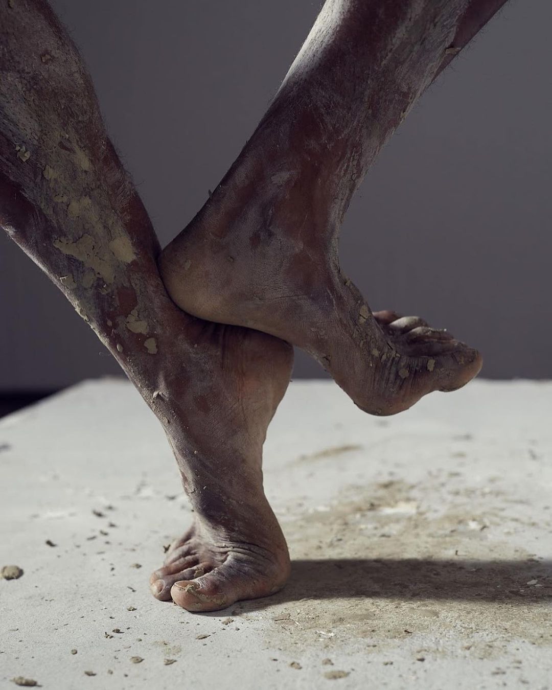 Close-up of dark-skinned adult legs covered in light-colored powder, intertwined on a concrete surface against a gray background.