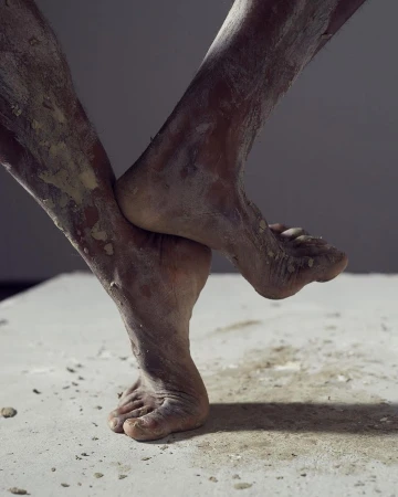 Close-up of dark-skinned adult legs covered in light-colored powder, intertwined on a concrete surface against a gray background.