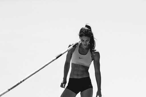 Athletic woman in sports bra and shorts, carrying a pole vault pole, with a clear white sky background; fitness and sport theme.
