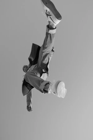 Model captured mid-air in a dynamic low-angle shot wearing cuffed jeans, jacket, and sneakers against a grey backdrop.