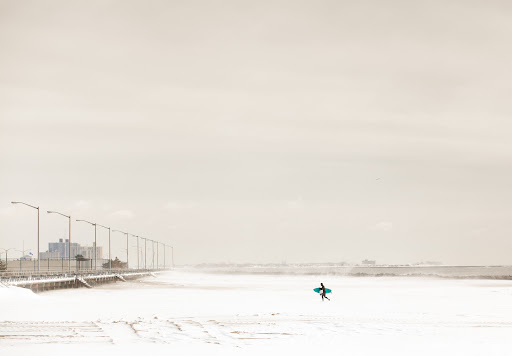 Lone surfer carrying surfboard walks along a snowy beach near a pier, under a bright overcast sky, creating a serene yet stark landscape.