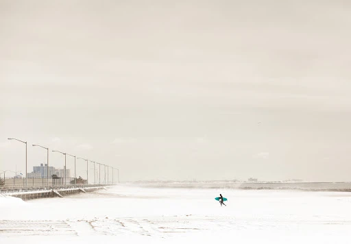 Lone surfer carrying surfboard walks along a snowy beach near a pier, under a bright overcast sky, creating a serene yet stark landscape.