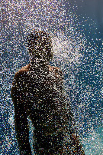 Man stands under a cascade of water against a blue sky, droplets create a shimmering effect, obscuring details.