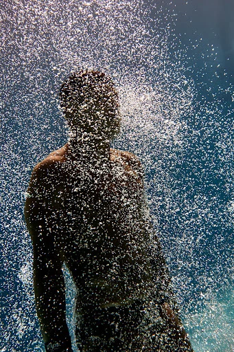 Man stands under a cascade of water against a blue sky, droplets create a shimmering effect, obscuring details.