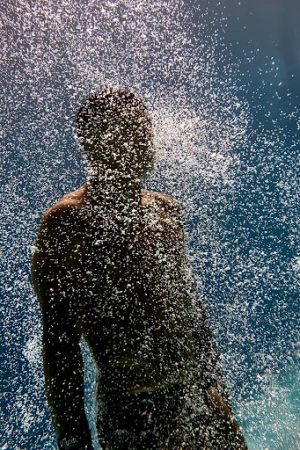 Man stands under a cascade of water against a blue sky, droplets create a shimmering effect, obscuring details.