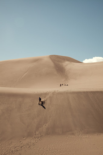 Person sandboarding down a sand dune in a desert landscape under a bright blue sky, with other people visible in the background.