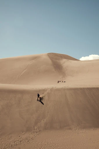 Person sandboarding down a sand dune in a desert landscape under a bright blue sky, with other people visible in the background.