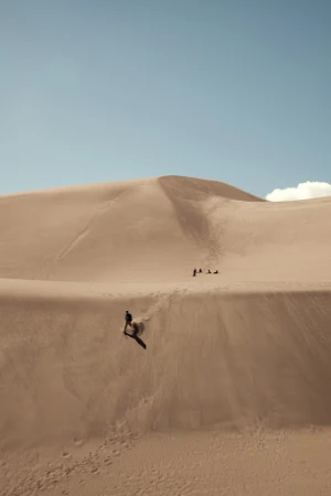 Person sandboarding down a sand dune in a desert landscape under a bright blue sky, with other people visible in the background.