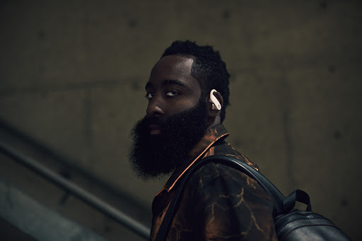 Close-up portrait of a black male athlete with a beard and backpack, wearing a stylish patterned jacket and wireless earbuds.