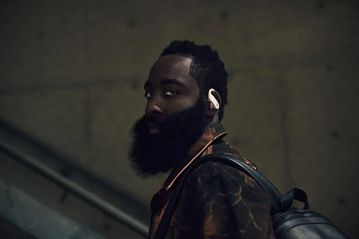 Close-up portrait of a black male athlete with a beard and backpack, wearing a stylish patterned jacket and wireless earbuds.