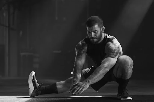 Monochrome shot of a muscular man stretching his groin in gym clothing in a dimly lit space with his hands on the floor.