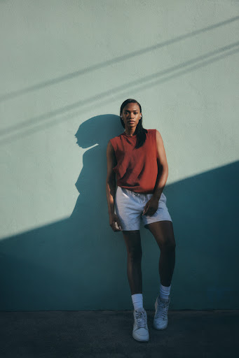 Woman poses outdoors wearing a rust-colored top, white shorts, white socks, and sneakers, leaning against a light blue wall.