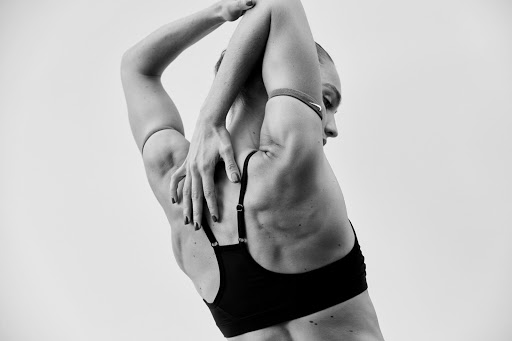 Black and white studio shot of a woman stretching her arms behind her back, wearing a black sports bra against a light background.