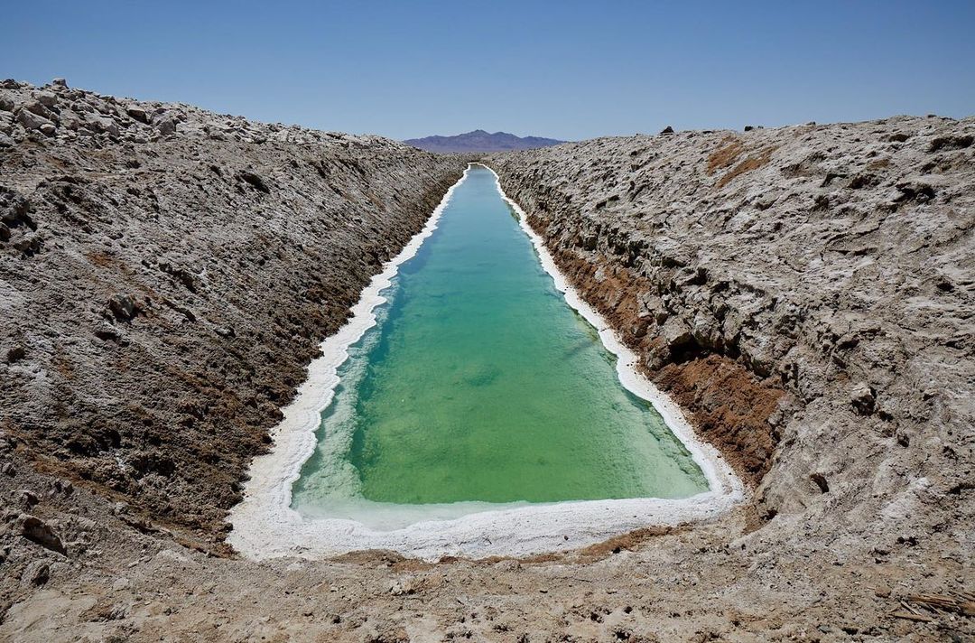 An eye-level shot showcases a long, narrow pool of turquoise water enclosed by earthen banks, under a clear blue sky. Mountain range in background.
