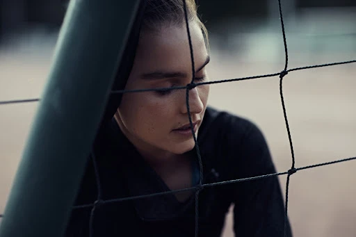 A young adult woman wearing a dark top is seen through a volleyball net in an outdoor setting, with a shallow depth of field.