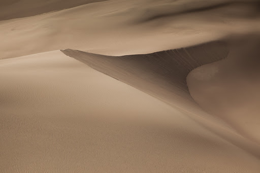 Abstract landscape featuring sand dunes with a gradient of light and shadow, showcasing natural desert patterns and textures in a monochromatic color scheme.