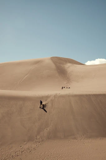 Person walking down large sand dune with clear blue sky in the background; several people visible in the distance.