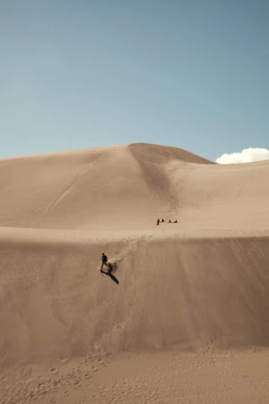 Person walking down large sand dune with clear blue sky in the background; several people visible in the distance.