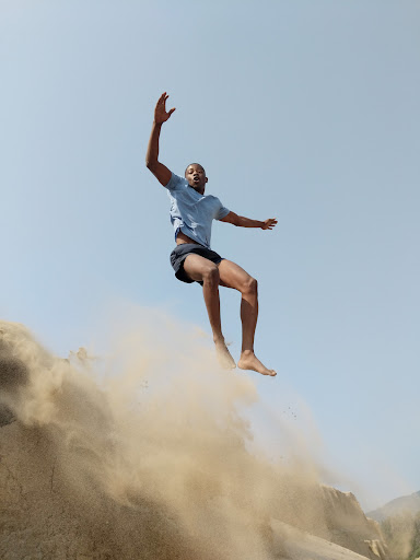 African-American man jumping from a sand dune against a blue sky. He's wearing a blue t-shirt and dark shorts in bright sunlight.