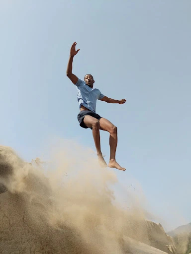 African-American man jumping from a sand dune against a blue sky. He's wearing a blue t-shirt and dark shorts in bright sunlight.