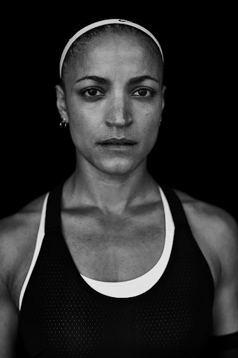 Black and white portrait of a determined woman wearing a sports bra and headband against a solid black background.