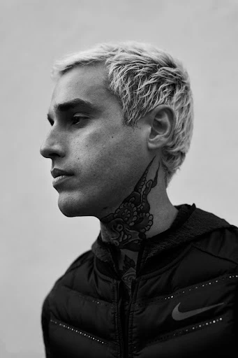 Black-and-white portrait of a young adult male with platinum blonde hair, a neck tattoo, and a black puffer jacket against a white background.