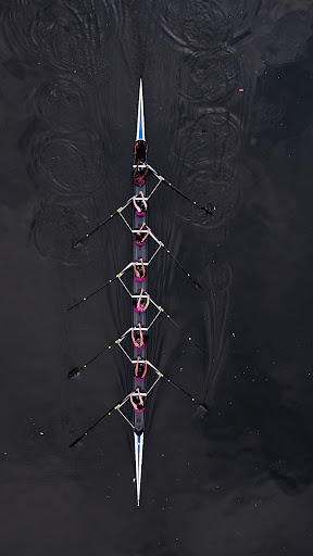 Aerial view of an eight-person rowing team with oars synchronized in dark water, creating ripples and symmetrical patterns during a race.