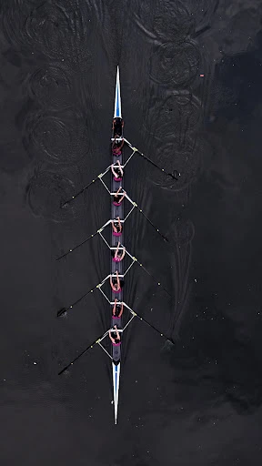 Aerial view of an eight-person rowing team with oars synchronized in dark water, creating ripples and symmetrical patterns during a race.