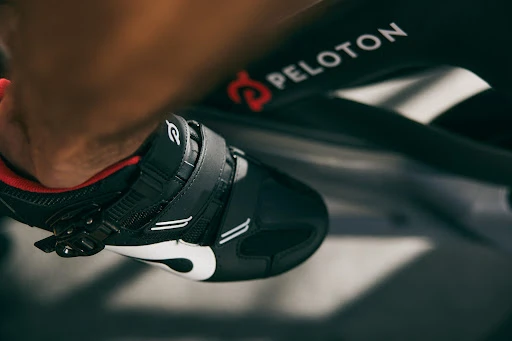 Close-up of a person's foot in a black and white Peloton cycling shoe clipped into the pedal of a Peloton exercise bike.