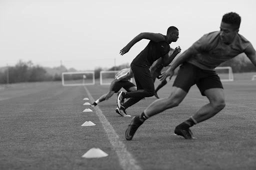 Black and white action shot of four male athletes sprinting on a grass field with white cones and a goalpost in the background.