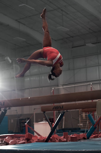 A female gymnast in mid-air performing a backflip over a balance beam in a gymnasium setting, showcasing athleticism and skill.