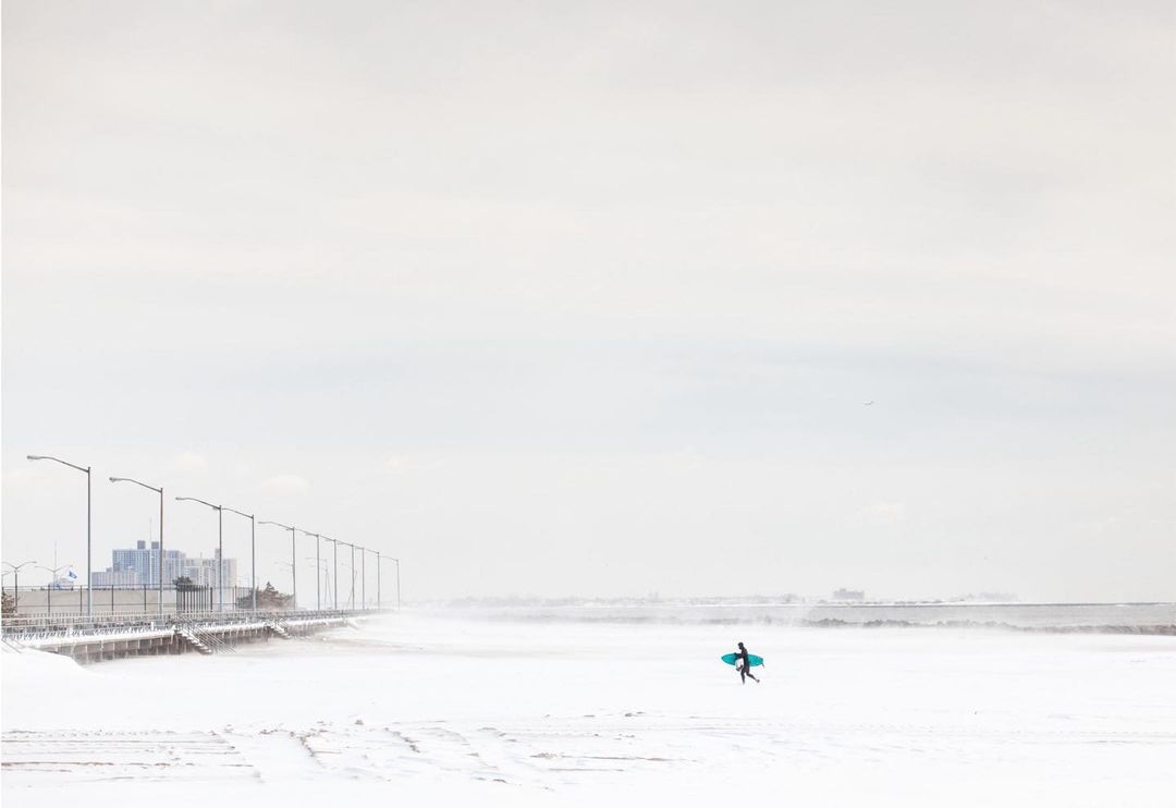 Lone surfer in wetsuit walks across snow-covered beach with teal surfboard, buildings visible in background against pale sky.