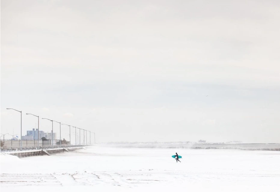 Lone surfer in wetsuit walks across snow-covered beach with teal surfboard, buildings visible in background against pale sky.