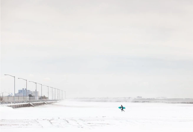 Lone surfer in wetsuit walks across snow-covered beach with teal surfboard, buildings visible in background against pale sky.