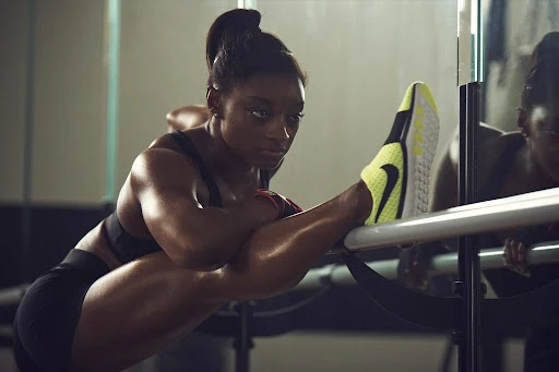Athlete Simone Biles stretches at a barre in a fitness studio. She wears black athletic apparel and yellow and black Nike training shoes.