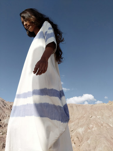 Woman in a long white dress with blue stripes standing against a dry desert landscape under a clear sky.
