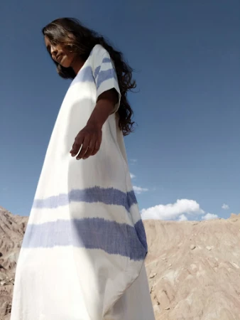 Woman in a long white dress with blue stripes standing against a dry desert landscape under a clear sky.