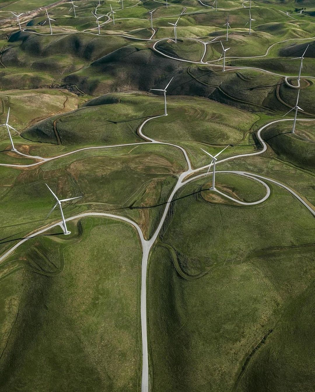 Aerial view of wind turbines on rolling green hills with white gravel paths winding through the landscape, generating clean energy.