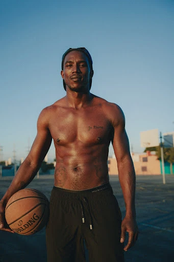 An athletic, shirtless African-American man holds a basketball outdoors against a blue sky, showcasing physical fitness and an active lifestyle.