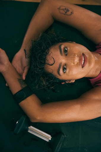A woman with tattoos lies on a mat with a dumbbell. Sweaty, dark curly hair, looking at the camera. Fitness and active lifestyle.