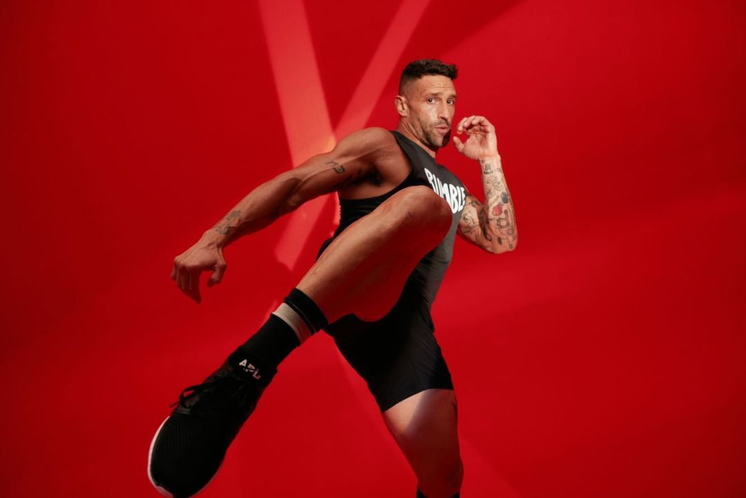Athletic man in black workout clothes demonstrates a kick against a red background with studio lighting and visible tattoos.